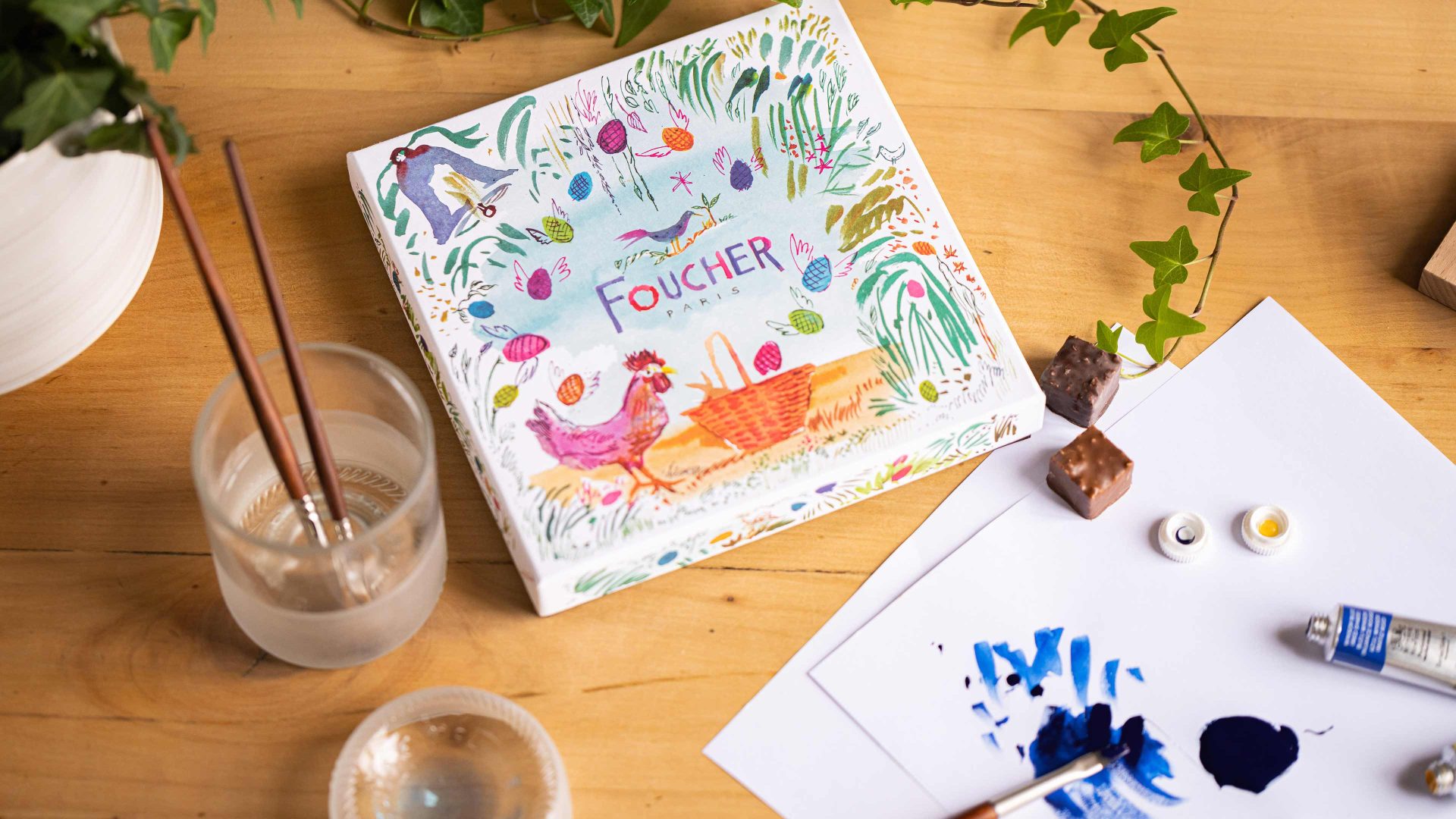 Une boîte Foucher entourée d'une plante, de pinceaux à tremper, de deux chocolats, ainsi que de feuilles blanches avec des traces d'aquarelles ainsi qu'un tube de peinture.