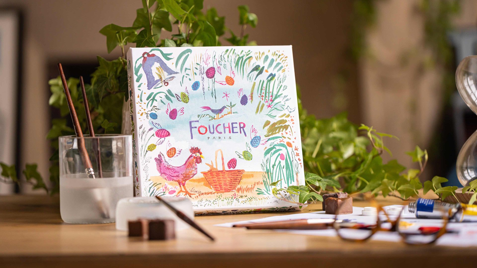 Une boîte Foucher avec son style aquarelle. La boîte est entourée de pinceaux et de chocolats ainsi que d'une plante;