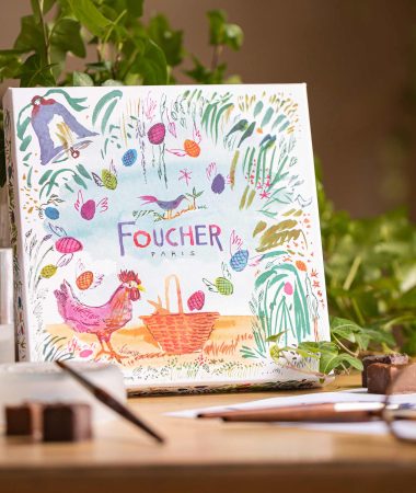 Une boîte Foucher avec son style aquarelle. La boîte est entourée de pinceaux et de chocolats ainsi que d'une plante;