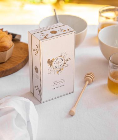 Une boîte de la Compagnie du Miel posée sur une table blanche. La boîte est entourée de bols, de pots de miels, de serviettes blanches, d'assiettes, d'une brioche ainsi que d'une cuillère à miel.
