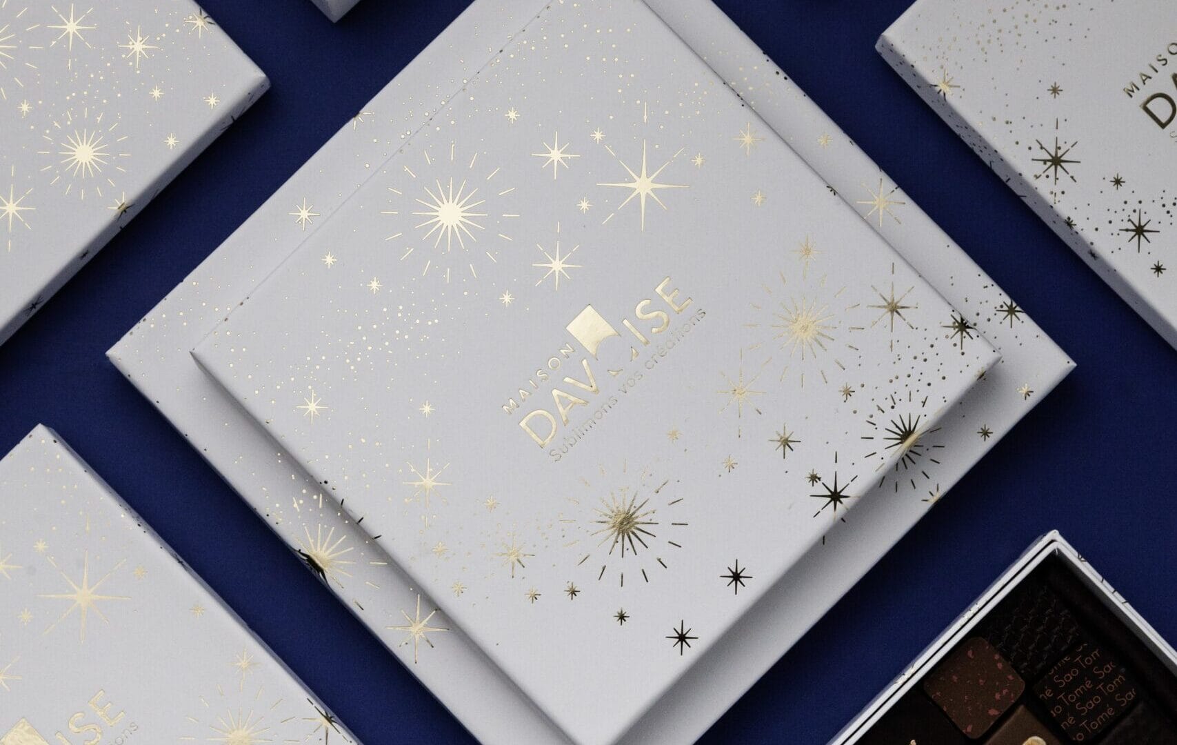 Assortiment de coffrets de chocolats blancs décorés d’étoiles dorées, disposés sur fond bleu, avec un coffret ouvert révélant des chocolats assortis.