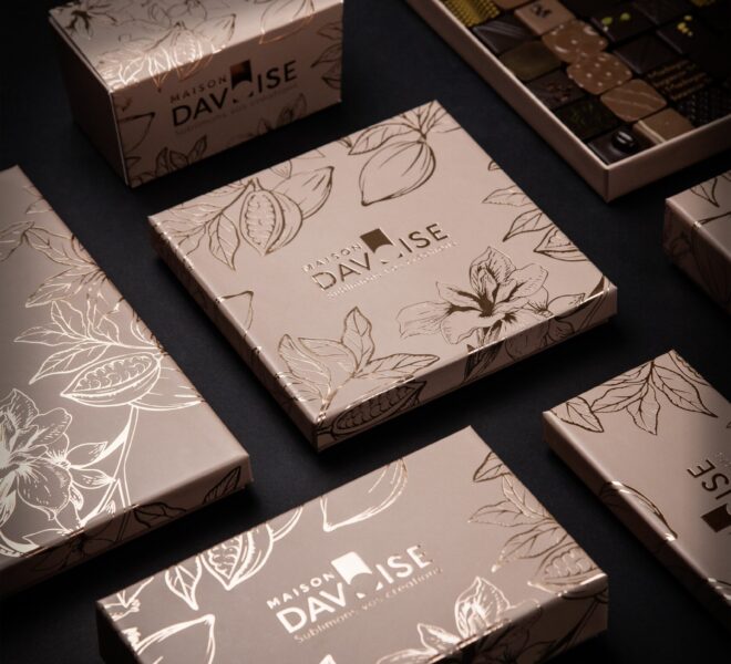 Coffrets de chocolats beige doré ornés de motifs floraux, posés sur fond sombre, avec un plateau de chocolats fins visible en arrière-plan.