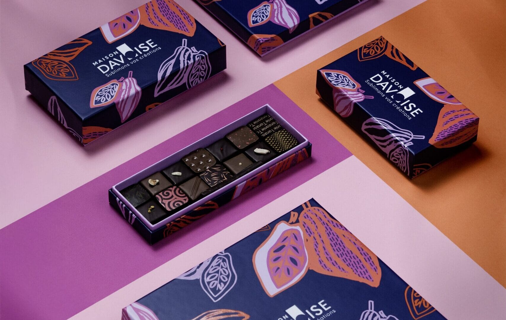 Boîtes de chocolats bleu nuit illustrées de cabosses colorées, présentées sur un fond rose et orange, avec un coffret ouvert montrant des chocolats variés.