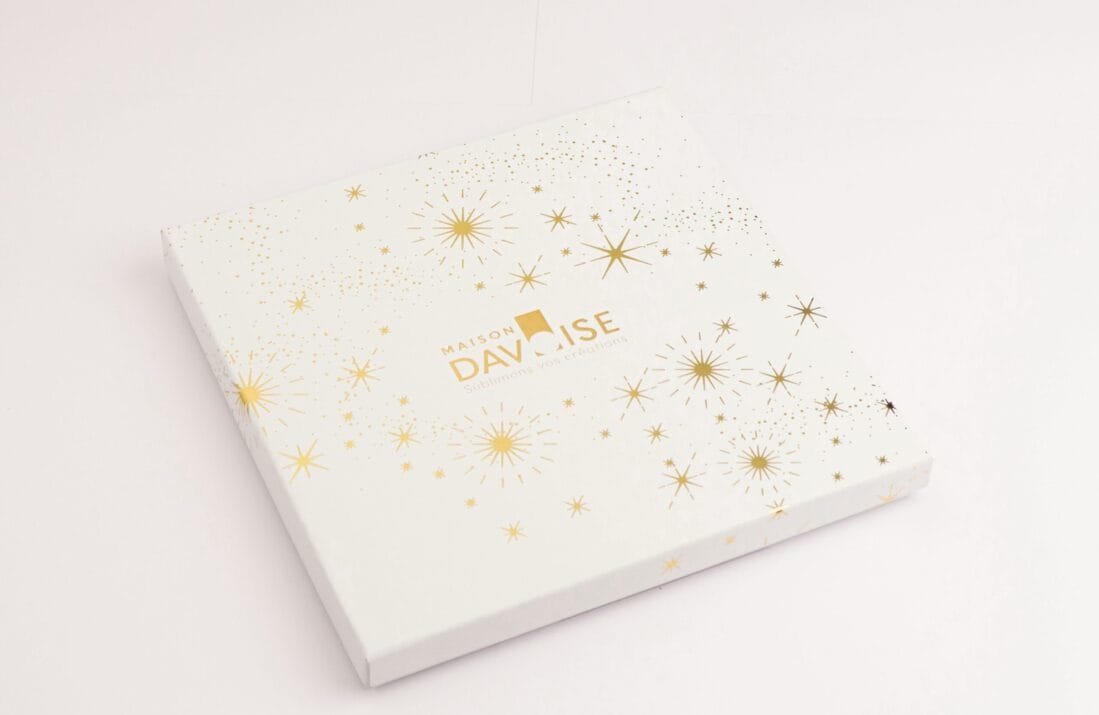 Boîte carrée Maison Davaïse blanche décorée d’étoiles dorées, fermée sur fond neutre.
